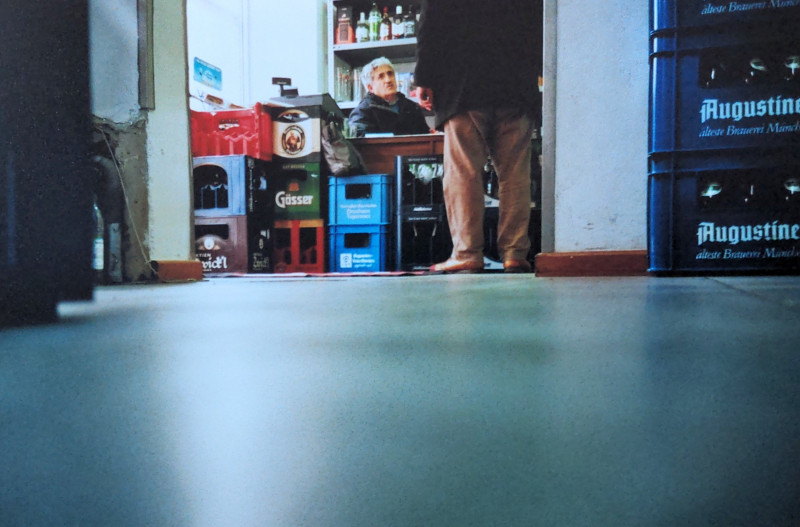 Ein analoges Farbfoto von Aenestis, wie er jemand bedient, es ist vom Boden aus fotografiert, zu sehen ist ein Kunde (nur die Beine), wie er mit Aenestis spricht.