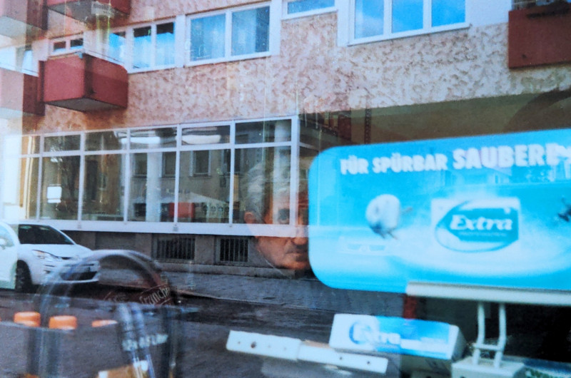 Ein analoges Farbfoto durch das Fenster der Getränkequelle fotografiert. Man sieht Ahmadi, wie er nach außen schaut. und die Balkone des Hauses gegenüber spiegeln sich im Fenster.
