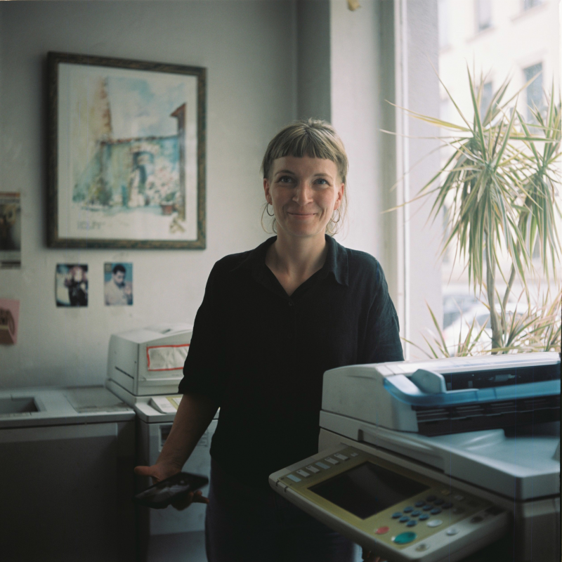 Miriam Worek (Frau mit kurzem Pony und schwarzem Oberteil) grinsend hinter einem Kopierer vor einem Fenster mit Palme (Kopierzentrum Luisenstraße)
